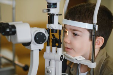 Genç bir çocuğun bir klinikte phoropter kullanan bir göz doktoru tarafından gözleri kontrol ediliyor.