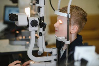 Genç bir çocuk, modern bir göz kliniğinde bir göz doktorunun rehberliğinde bir phoropter kullanılarak sabırla görme testinden geçiyor.