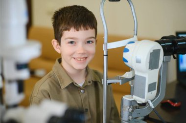 Modern bir göz hastalıkları kliniğinde ileri seviye teşhis cihazları kullanarak göz muayenesi sırasında gülümseyen genç çocuk, çocukların göz sağlığını ön plana çıkarıyor.