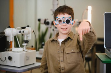 Deneme çerçevesi takan gülümseyen çocuk bir göz hekimliği kliniğindeki görüş testinde göz muayenesi sürecini gösteriyor.