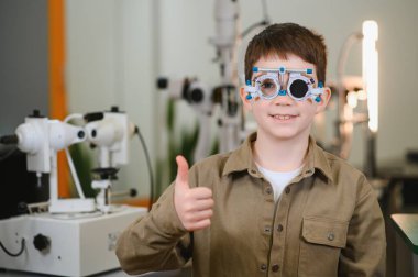 Bir göz hekimliği kliniğinde, sağlık sigortası konseptinde görme testinde baş parmağını gösteren bir deneme çerçevesi takan genç bir çocuk.