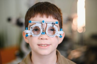 Göz muayenesi sırasında göz doktorunun muayenehanesinde deneme çerçevesi takan gülümseyen çocuk, görüş bakımına neşeli bir yaklaşım sergiliyor.