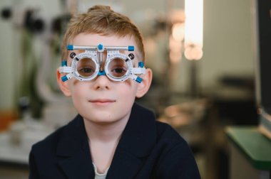 Deneme çerçevesi takan genç bir çocuk göz doktorunun ofisinde göz muayenesinden geçiyor, görüş değerlendirmesi ve bakıma odaklanıyor.