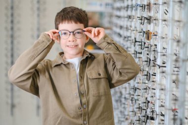 Çocuk optik mağazada yeni gözlüklerini ayarlıyor, gülümsüyor ve seçiminden memnun görünüyor.