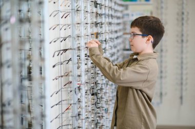 Bir optik mağazasında çeşitli gözlük deneyen çocuk, optik görme düzeltmesi ve göz sağlığını geliştirmek için çerçeveler seçiyor