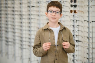 Optik mağazasında yeni gözlük deneyen çocuk, görme düzeltmesi ve sağlık hizmetleri ile göz doktorundan yardım alıyor.