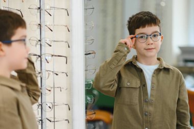 Görme tedavisi sırasında göz hekimliği kliniğinde aynanın önünde yeni gözlük takan çocuk.