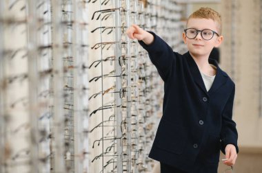 Bir optik mağazada sergilenen gözlüklere işaret eden çocuk, bir gözlükçünün yardımıyla yeni gözlük seçiyor.