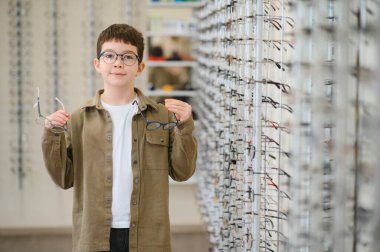 Bir optik mağazada gözlük takan çocuk, görüş düzeltmesi için farklı çerçeveler keşfediyor, pediatrik göz bakımının önemini gösteriyor.