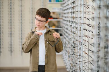 Bir optik mağazada yeni gözlük deneyen çocuk, görme ve göz sağlığını geliştirmek için çeşitli çerçeveler keşfediyor.