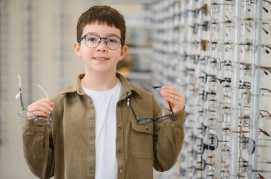 Gözlüklü, gülümseyen çocuk bir gözlük dükkanında iki gözlük tutuyor ve görüş düzeltmesi için en iyisini seçiyor.