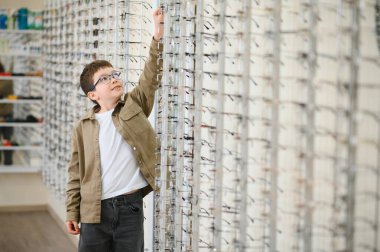 Gözlüklü çocuk bir optik mağazada yeni bir gözlüğe uzanıyor, görüş düzeltme seçeneklerini keşfediyor.