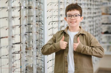 Mutlu çocuk yeni gözlük takıyor, başparmağını kaldırıyor ve çeşitli gözlük seçenekleriyle dolu bir optik mağazada gururla duruyor.