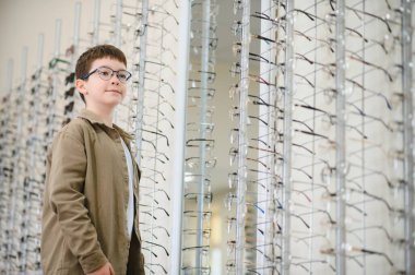 Gözlük takan bir çocuk bir optik mağazada gözlük çerçevesinin yanında duruyor ve yeni gözlükler seçiyor.