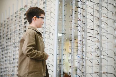 Gözlüklü genç, optik mağazada farklı çerçeveler deneyerek yeni bir gözlük seçerken gülümsüyor.