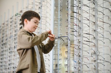 Genç bir çocuk, bir optik mağazadaki gözlüklerden dikkatlice gözlük seçiyor ve çocuklar için vizyon bakımının önemini vurguluyor.