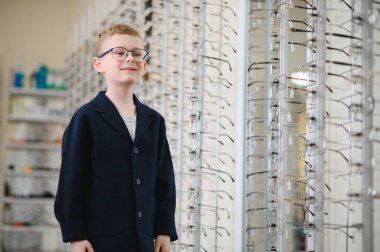 Çocuk yeni gözlük deniyor, göz doktorunun optik mağazayı ziyaretinden sonra net görüşün tadını çıkarıyor, göz seçimiyle mutlu.