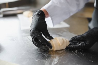 Siyah eldivenler giyen fırıncı profesyonel mutfakta paslanmaz çelik masada kruvasan hamuru hazırlıyor.