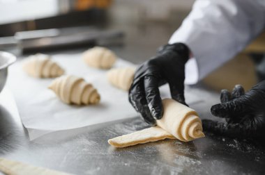 Siyah eldiven giyen fırıncı, hamuru paslanmaz çelik masaya ustalıkla sarar ve profesyonel mutfakta taze kruvasan pişirmeye hazırlanır.