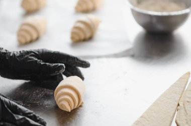 Fırıncı siyah eldiven giyip kruvasan hazırlıyor, pastanedeki paslanmaz çelik masaya taze rulo hamurlar koyuyor.