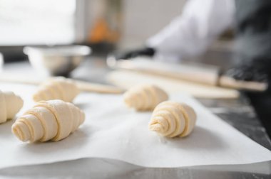 Pişirme kağıdındaki çiğ kruvasan yakın plan, profesyonel bir mutfağın arka planında çalışan bir fırıncı
