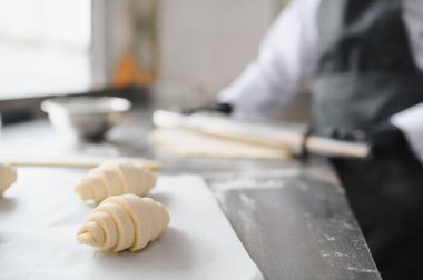 Parşömen kağıdındaki ham kruvasanların yakın plan çekimi. Profesyonel bir mutfağın arka planında çalışan bir fırıncı var.