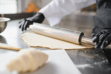 Fırıncı merdane kullanıyor, hamuru paslanmaz çelik masada düzleştiriyor, profesyonel mutfakta hamur işleri hazırlıyor.