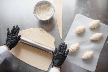 Fırıncı hamuru yassılaştırmak için merdane kullanıyor, yeni hazırlanmış kruvasanlarla, profesyonel bir mutfakta hamur işi yapma sürecini gösteriyor.