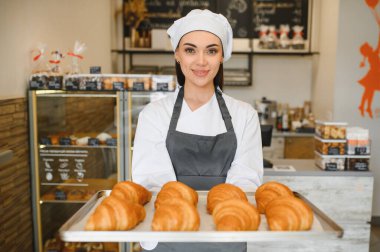 Bir tepsi altın rengi kruvasan sunan dişi fırıncı, fırın ortamında pişirme yeteneğini sergiliyor.