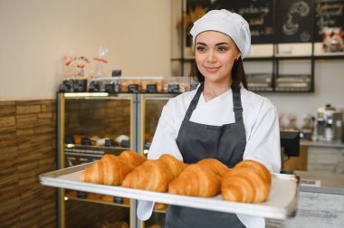Gülümseyen fırıncı kadın büyüleyici fırınında içi altın sarısı kruvasanlarla dolu bir tepsi sergiliyor. Sıcak ve lezzetli aromalar yayıyor.