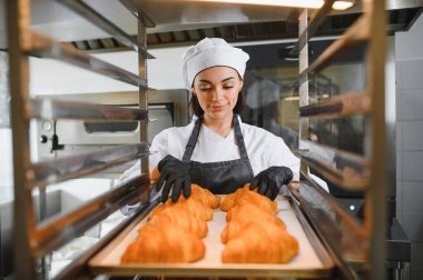 Siyah eldiven giyen kadın fırıncı profesyonel mutfakta tepside taze pişmiş altın kruvasan düzenliyor.