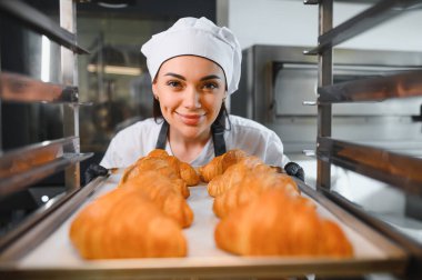 Siyah eldiven giyen kadın fırıncı ticari fırının profesyonel fırınına bir tepsi altın rengi kruvasan koyuyor.
