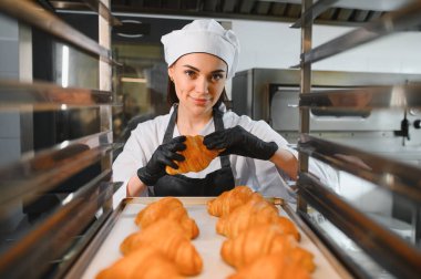 Profesyonel bir fırıncı, mükemmel bir kruvasanı gururla sergiliyor. Etrafı, ticari bir mutfakta bir tepsi altın hamur işi ile çevrili.