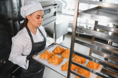 Profesyonel fırıncı gururla bir tepsi altın-kahverengi kruvasan sergiliyor. Ticari bir mutfakta pişirme sanatını ve adanmışlığını sergiliyor.