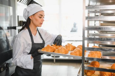 Profesyonel bir fırıncı, altın sarısı kruvasanlarla dolu bir tepsiyi hareketli bir ticari mutfağın rafına yerleştiriyor ve mutfak becerilerini sergiliyor.