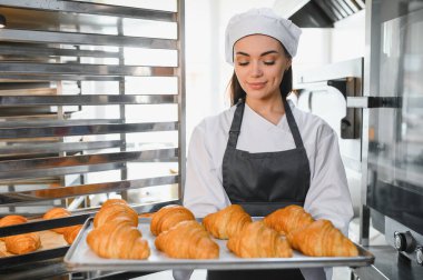 Profesyonel fırıncı gururla bir tepsi altın kruvasan sunar. Ticari bir mutfak ortamında pişirmenin sanatını ve adanmışlığını gözler önüne serer.