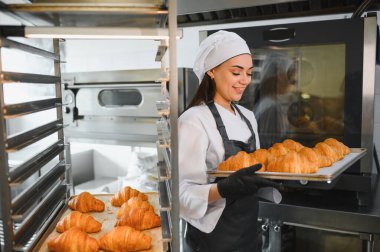 Elinde bir tepsi altın rengi kruvasan tutarken gülümseyen profesyonel fırıncı, ticari bir mutfak ortamında pişirme uzmanlığının sonucunu sergiliyor.