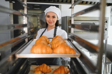 Profesyonel fırıncı gülümserken ticari bir fırındaki büyük bir endüstriyel fırına bir tepsi kruvasan yerleştiriyor.