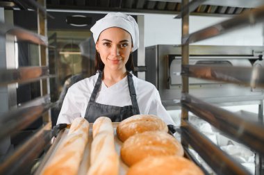 Bir tepsi taze ekmek ve baget sunan profesyonel fırıncı, bir fırın ortamında çalışmalarının sonucunu gözler önüne seriyor.