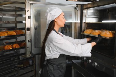 Profesyonel fırıncı siyah eldiven takıyor. Endüstriyel fırından altın kahverengi somun tepsisini çıkarıyor. Etrafı hamur işleriyle çevrili.