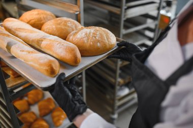Fırıncı siyah eldiven giyer, taze pişmiş ekmek tepsisi koyar ve ticari mutfakta raflara yuvarlak somunlar koyar.