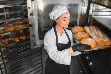 Profesyonel fırıncı, siyah eldiven ve önlük giyip endüstriyel fırına taze ekmek tepsisi koyuyor.