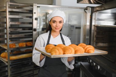 Profesyonel bir fırıncı, bir tepsi altın kruvasan sunuyor. Ticari bir mutfak ortamında pişirmenin sanatını ve adanmışlığını gözler önüne seriyor.