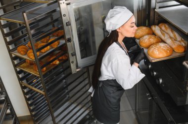 Profesyonel fırıncı, büyük bir fırından taze pişmiş bir tepsi ekmek alıyor. Yanında da kruvasan var.