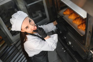 Profesyonel fırıncı, yeni pişmiş kruvasanları ticari bir mutfakta fırından çıkarırken dilini dışarı çıkarıyor.