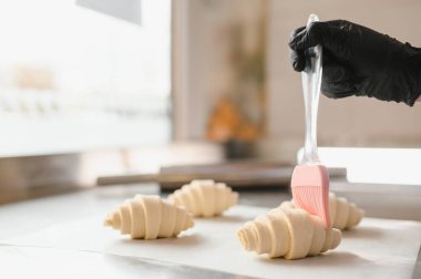 Siyah eldiven giyen fırıncı silikon fırçayla taze kruvasan fırçalayan profesyonel fırın mutfağında hamur işleri hazırlıyor.