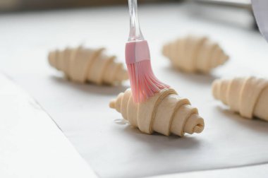 Fırıncı kruvasanları yumurta yıkama ile parlatıyor silikon fırça kullanıyor, profesyonel mutfakta pişirmeye hazırlanıyor.