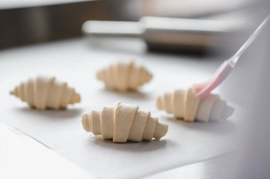 Fırınlanmamış kruvasanların yakın plan çekimleri. Parşömen kağıdında yumurta yıkama ile fırçalanıyorlar. Parlak bir mutfakta arka planda merdane var.