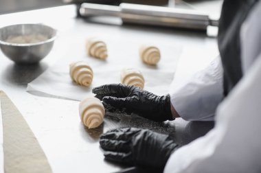 Fırında kruvasan hazırlayan siyah eldivenler giyen fırıncı onları paslanmaz çelik bir masaya yerleştiriyor.