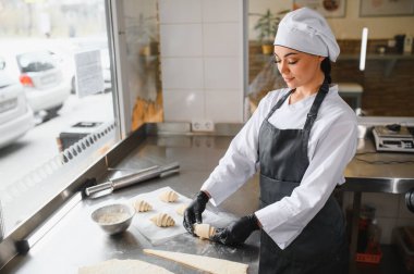 Siyah eldiven giyen kadın fırıncı taze hamur işlerinin zengin aromasıyla çevrili fırın mutfağında kruvasanları ustaca hazırlıyor.